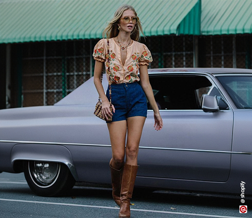 A woman walking in orange floral top, blue shorts and knee high boots
